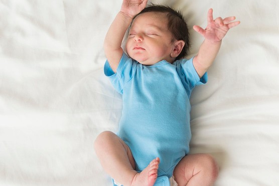 newborn baby boy lying on bed in a blue suit with arms in the air