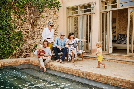 Wide shot of smiling grandparents relaxing with children and grandchildren beside pool at tropical villa during multigenerational family vacation